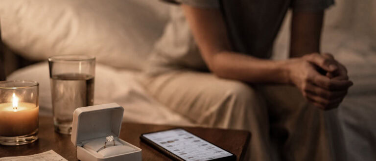 A woman sitting with a diamond ring in a box, next to a smartphone and two glasses of water on a wooden table.